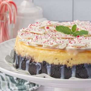 Side view of peppermint bark cheesecake with an Oreo crust, glossy white chocolate ganache drips, and pieces of peppermint bark layered on top, displayed on a white cake stand for a festive holiday dessert.