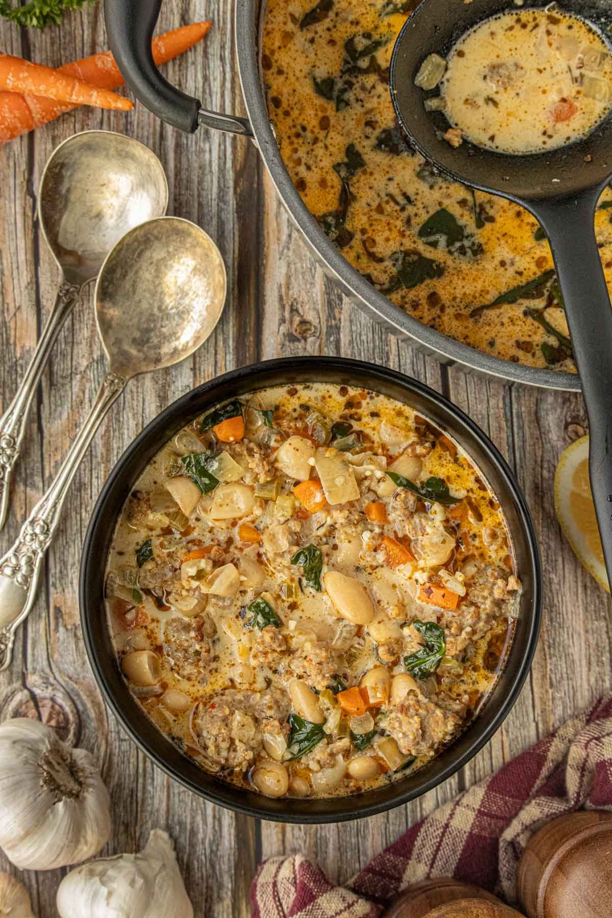 Bowl of creamy sausage and white bean soup with Italian sausage, white beans, carrots, and leafy greens on a rustic wooden table with bread and garlic. Photographed by Kim Ledet for Marye Audet at Restless Chipotle.