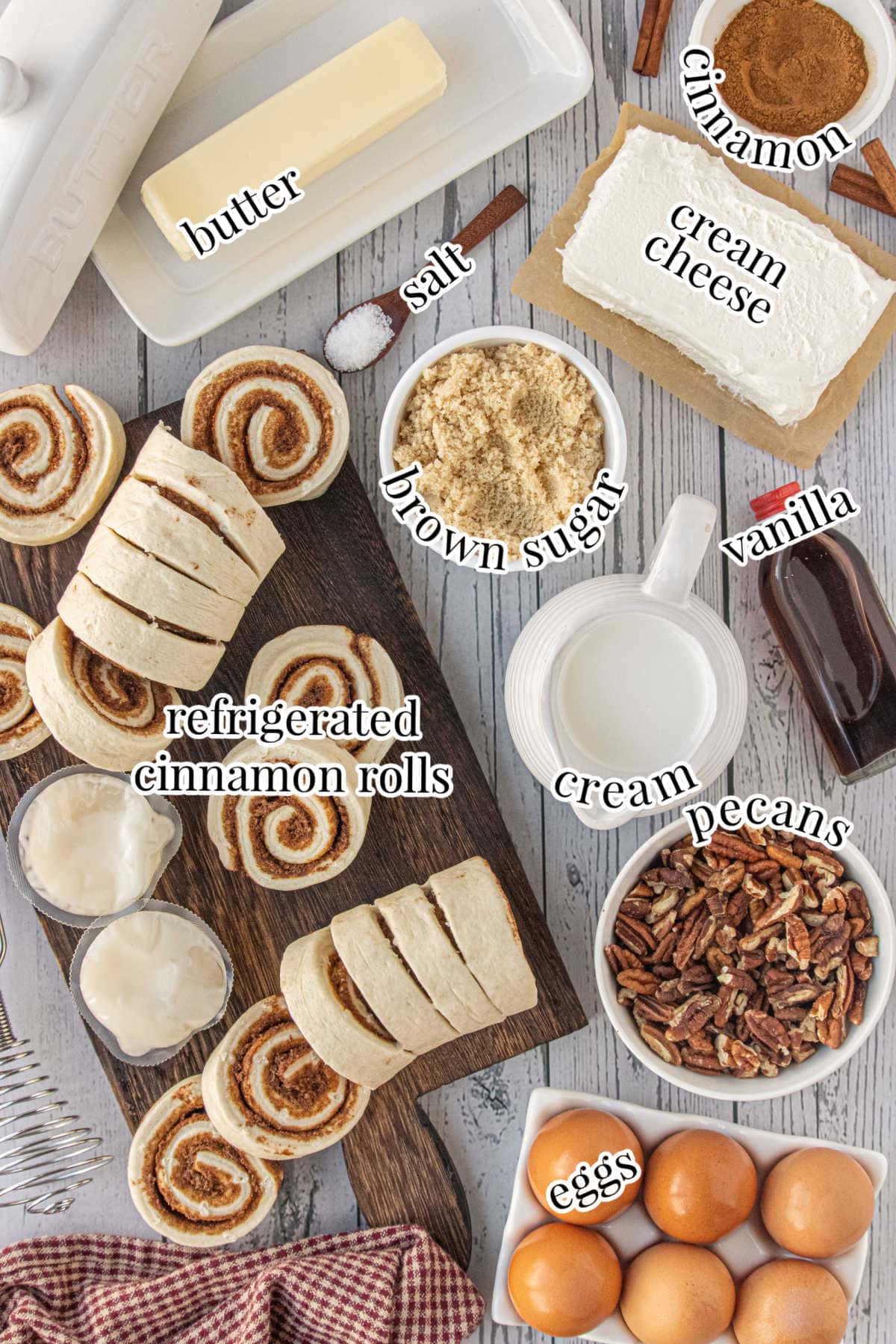 Overhead photo showing all the ingredients for cinnamon roll breakfast casserole, including sliced refrigerated cinnamon rolls, cream cheese, brown sugar, pecans, eggs, butter, cinnamon, vanilla, salt, and cream arranged on a wooden board and kitchen counter.