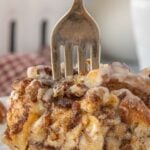 A close-up vertical Pinterest-style image showing a gooey slice of cinnamon roll breakfast casserole with icing and pecans, held in place by a fork. Text overlay reads “Domestic Goddess? Accidentally.” and “Cinnamon Roll Casserole.”
