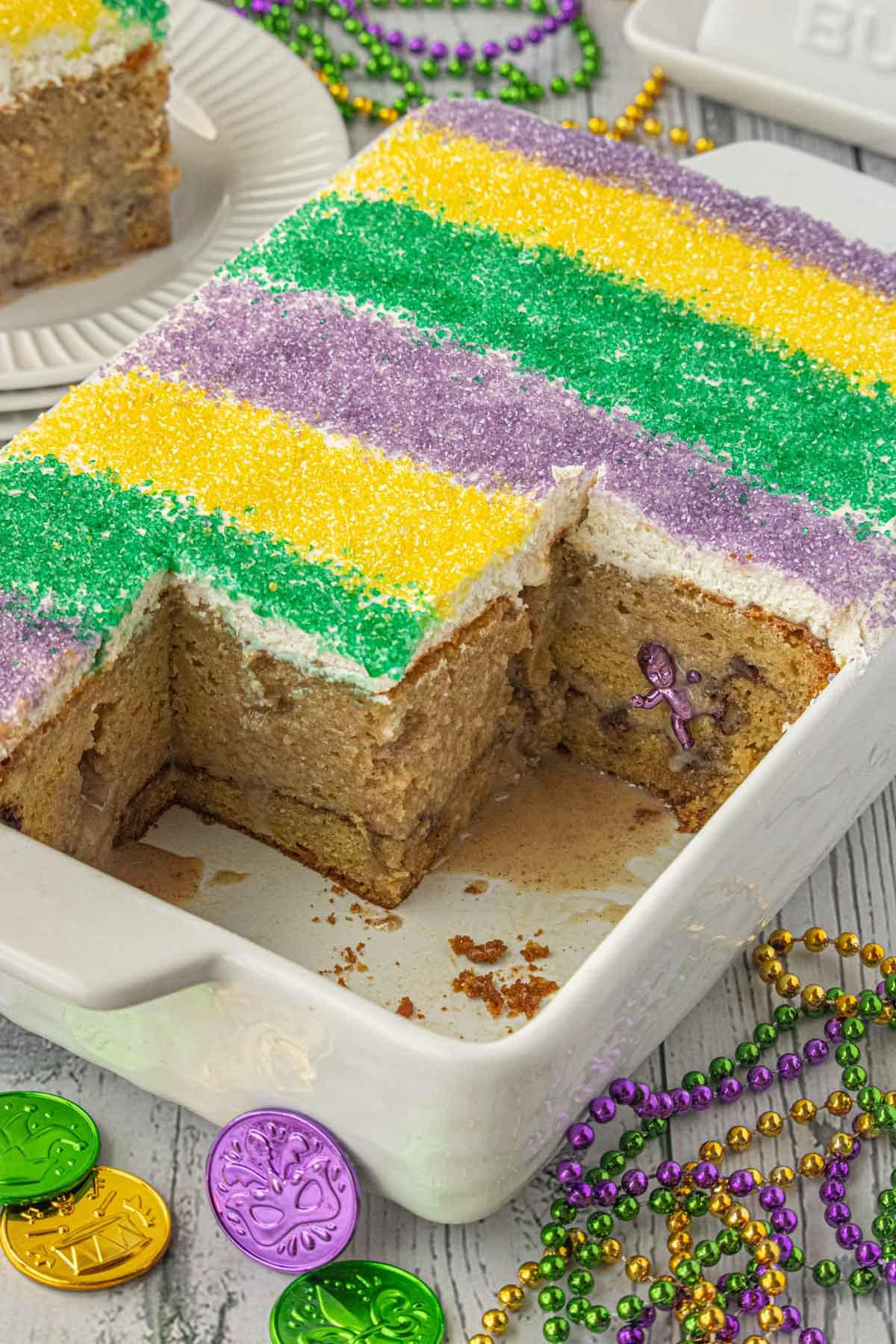 King cake poke cake in a white baking dish topped with purple, green, and gold sugar, with a slice removed showing cinnamon layers and a hidden plastic baby