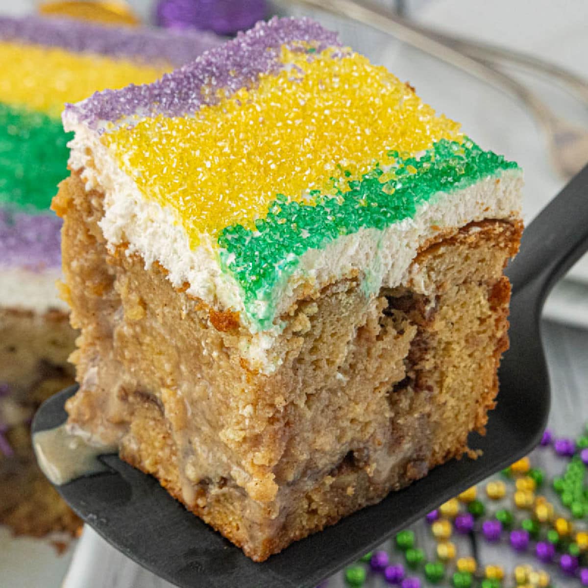 Close-up of king cake poke cake slice with cinnamon swirl layers and cream cheese frosting topped with purple, green, and gold sugar