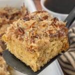 Close-up of old-fashioned oatmeal cake with coconut pecan topping on a spatula, with coffee in the background