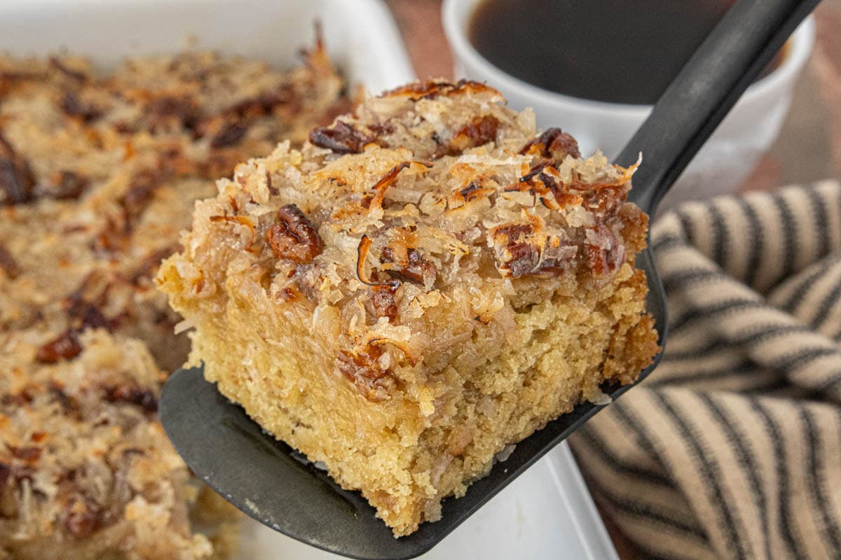 Close-up of old-fashioned oatmeal cake with coconut pecan topping on a spatula, with coffee in the background