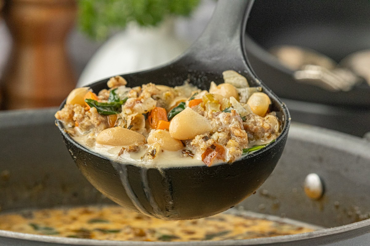 Ladle of creamy Italian sausage and cannellini bean soup with sausage, white beans, carrots, and greens held over a pot of simmering soup, photographed by Kim Ledet for Marye at Restless Chipotle.