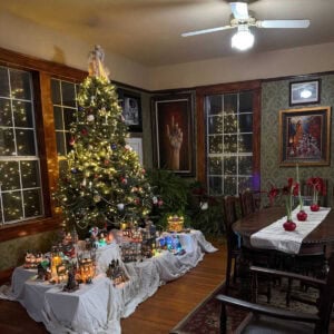 A cozy dining room decorated for Christmas with a lit tree, a vintage ceramic Christmas village displayed beneath it, twinkling lights in the windows, and a wooden dining table set with red candles.