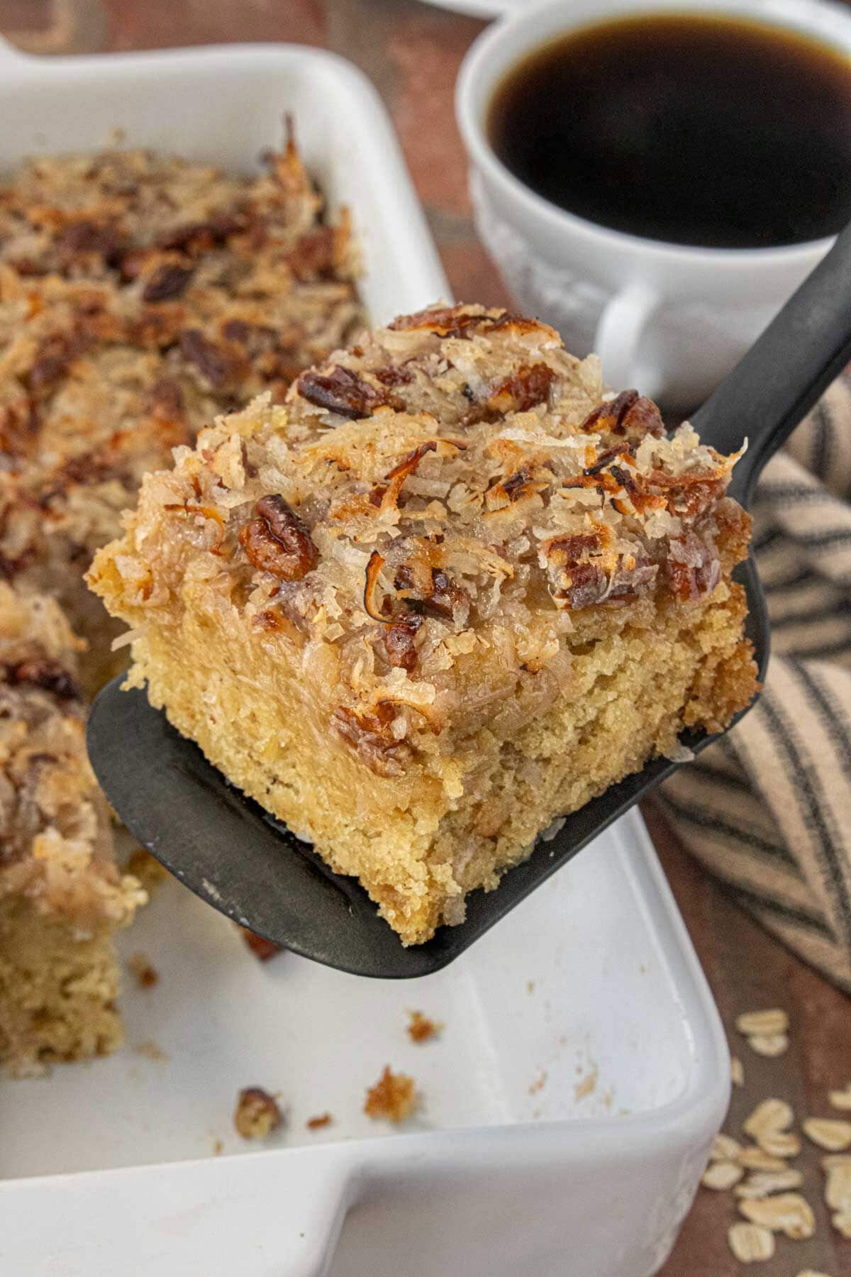 Close-up of old-fashioned oatmeal cake with coconut pecan topping on a black spatula, with a cup of coffee in the background