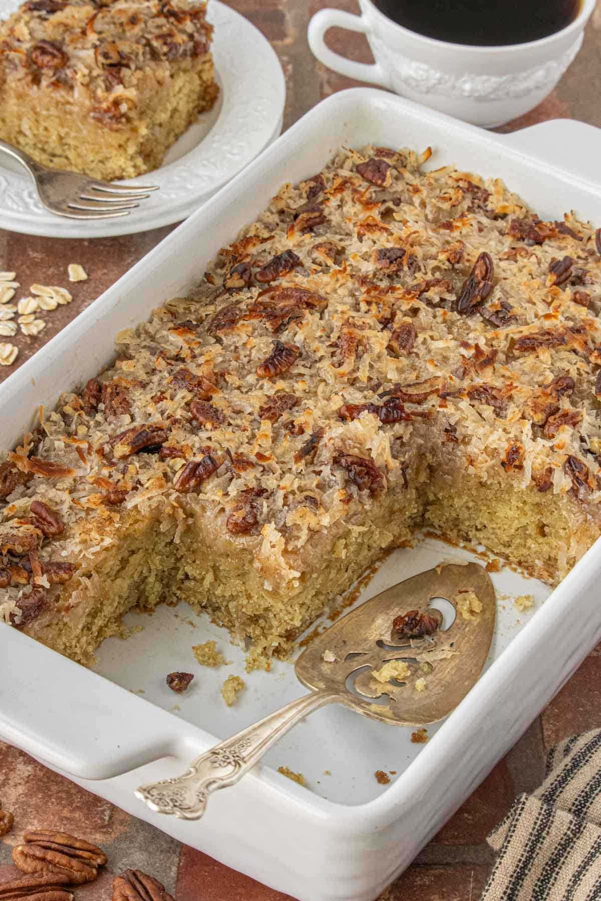 Old-fashioned oatmeal cake baked in a white casserole dish with a scoop removed, showing the moist crumb and coconut pecan topping