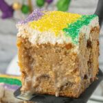 King cake poke cake slice on a spatula with cinnamon layers, condensed milk soak, cream cheese frosting, and purple, green, and gold Mardi Gras sugar, with text reading “Still counts as king cake”