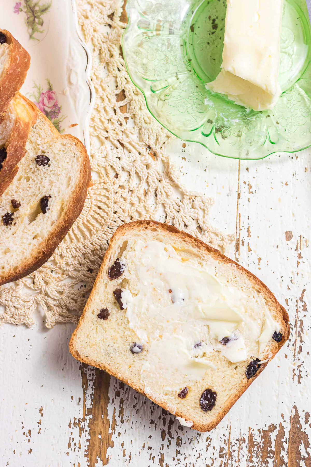 Slice of homemade raisin bread spread with butter on rustic white table with green glass dish