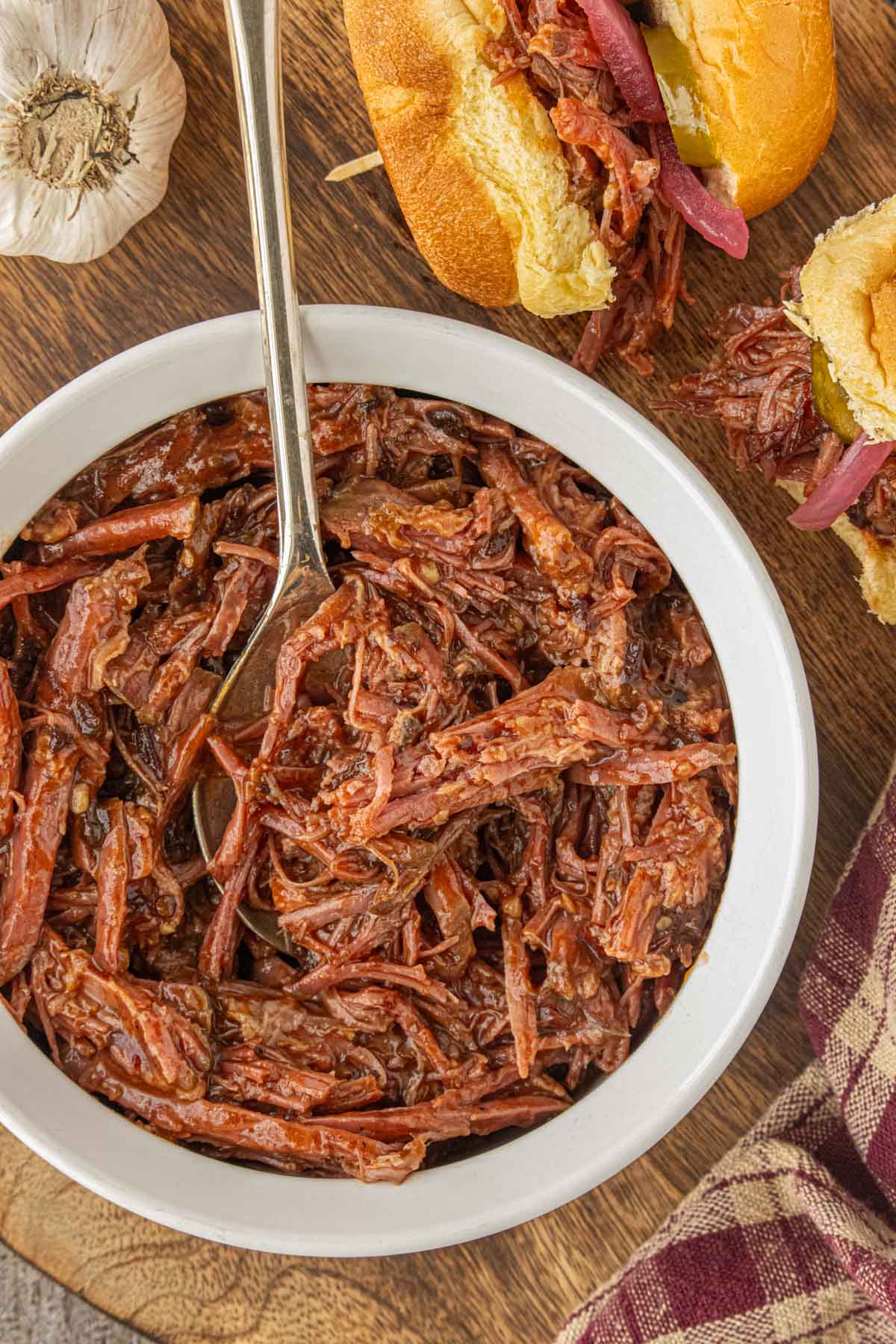 Slow cooker shredded brisket in barbecue sauce served in a white bowl with slider buns, pickles, and pickled onions on a wooden board.
