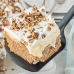 Carrot cake poke cake slice on white plate with thick cheesecake pudding layer, whipped topping, and pecans.