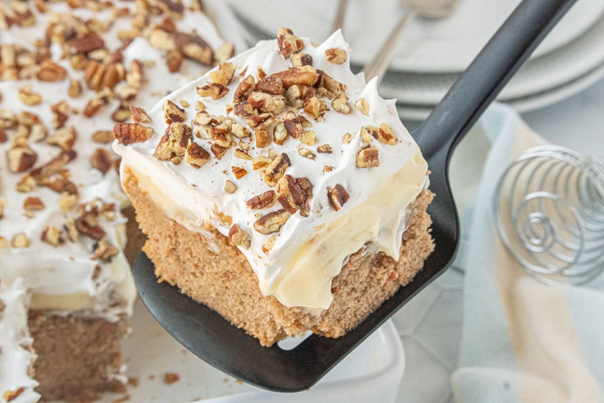 Carrot cake poke cake slice on white plate with thick cheesecake pudding layer, whipped topping, and pecans.