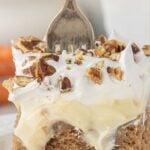Close-up slice of carrot cake poke cake topped with cheesecake pudding layer, whipped topping, and chopped pecans, with fork pressed into the cake. Title text overlay for Pinterest.