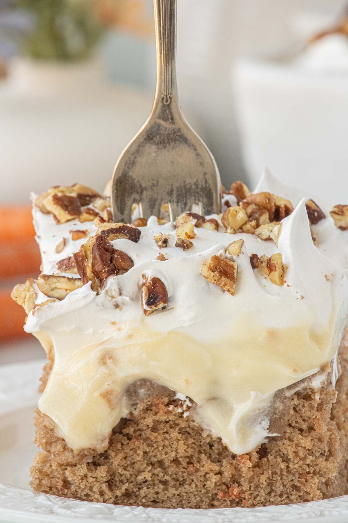 Close-up of carrot cake poke cake slice with creamy cheesecake pudding filling, whipped topping, and pecans, fork pressed into the top.