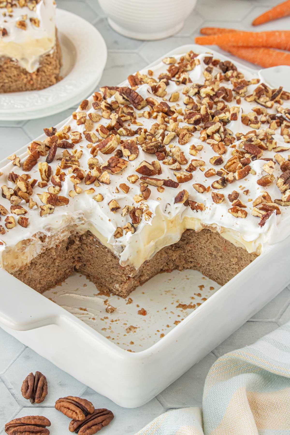 Carrot cake poke cake in a white baking dish topped with cream cheese pudding, whipped topping, and chopped pecans, with one large slice removed.