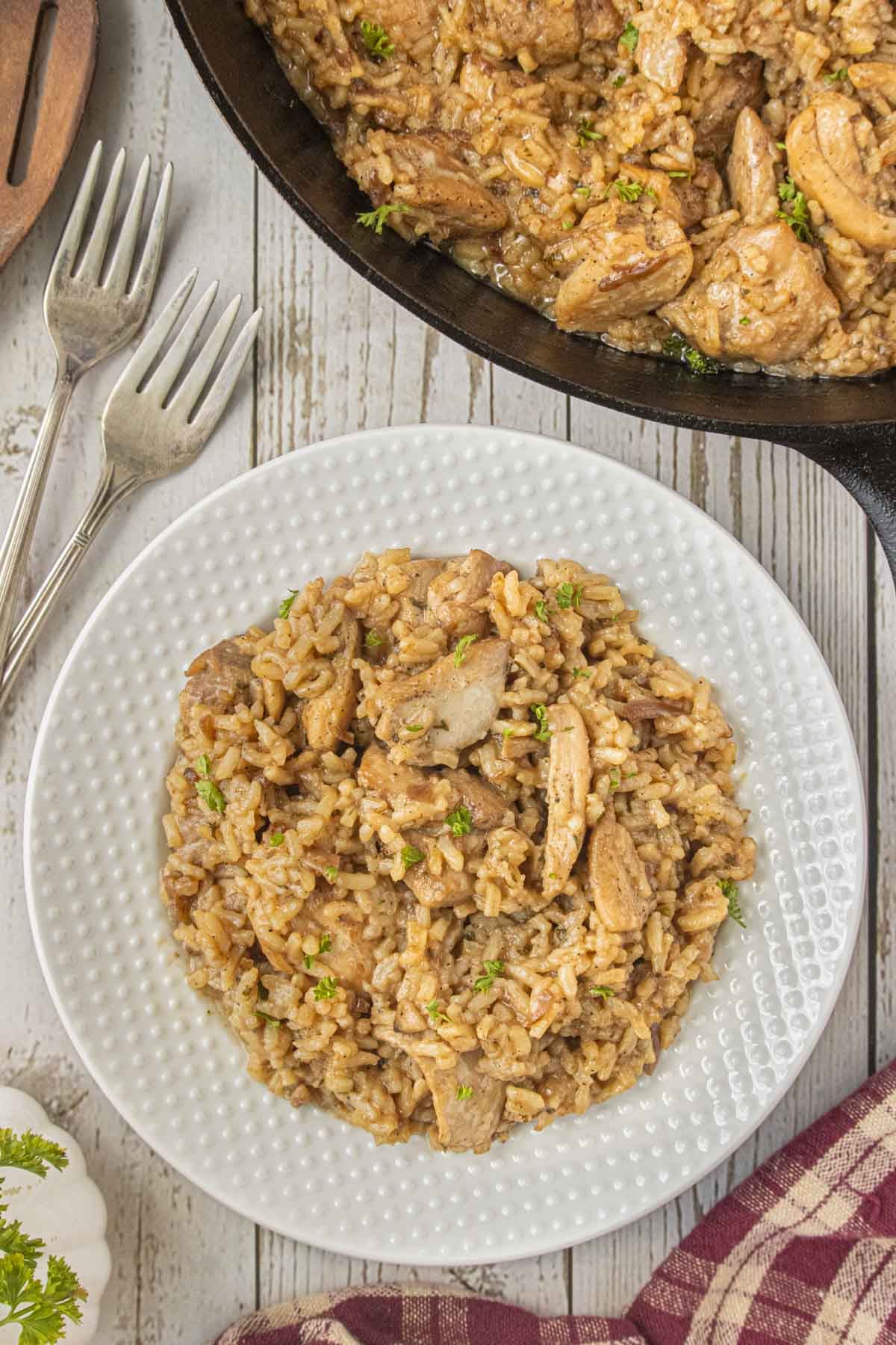 Serving of stick of butter chicken and rice casserole on a white plate with additional casserole in a cast iron skillet in the background.