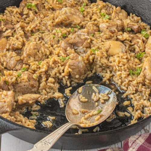Stick of butter chicken and rice casserole in a cast iron skillet with tender chicken, seasoned rice, and a serving spoon.