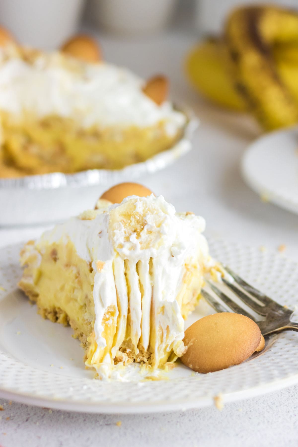 Slice of banana pudding pie with whipped topping, Nilla Wafers, and graham cracker crust on a white dessert plate.