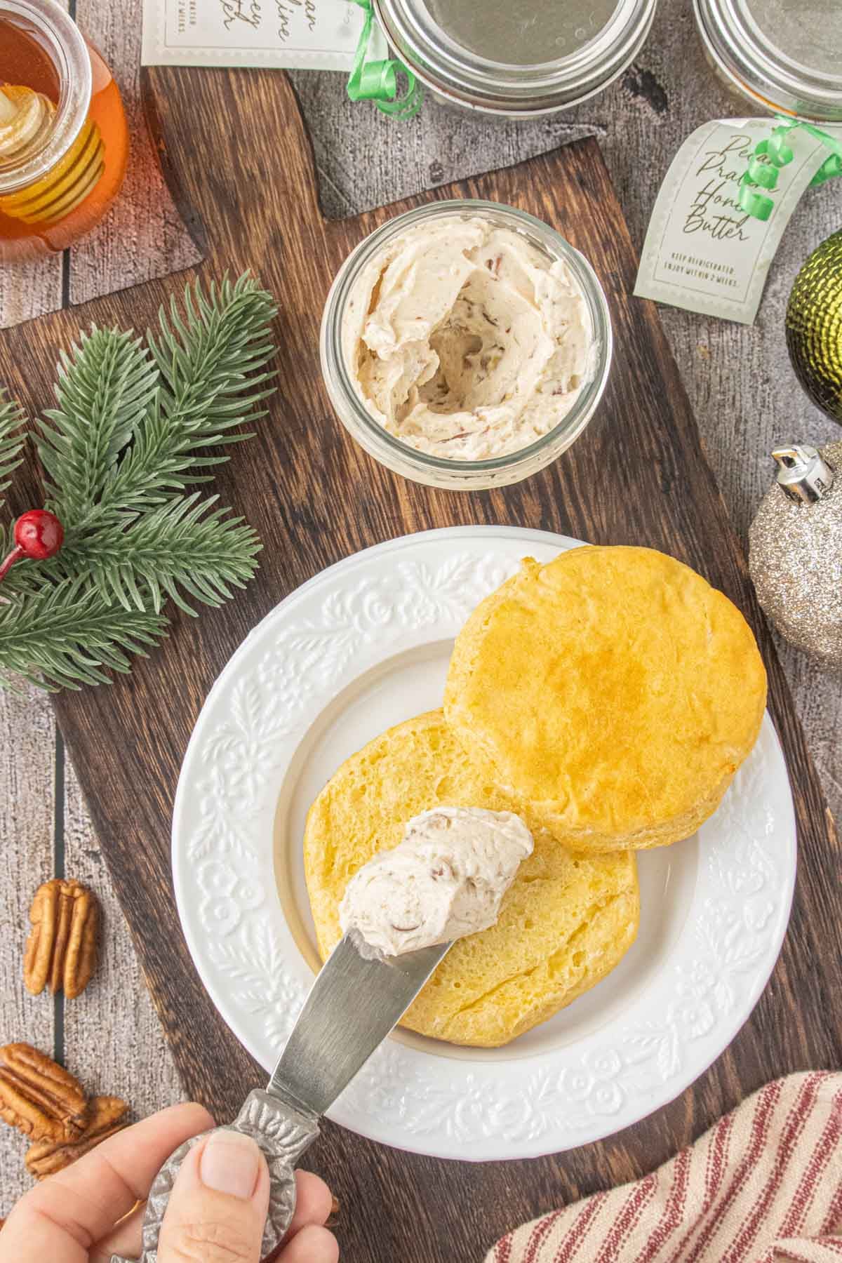 Spreading pecan praline honey butter onto a warm homemade biscuit with extra jars of flavored butter in the background.