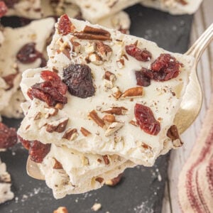 White chocolate cranberry bark stacked on a serving spoon with dried cranberries and chopped pecans.