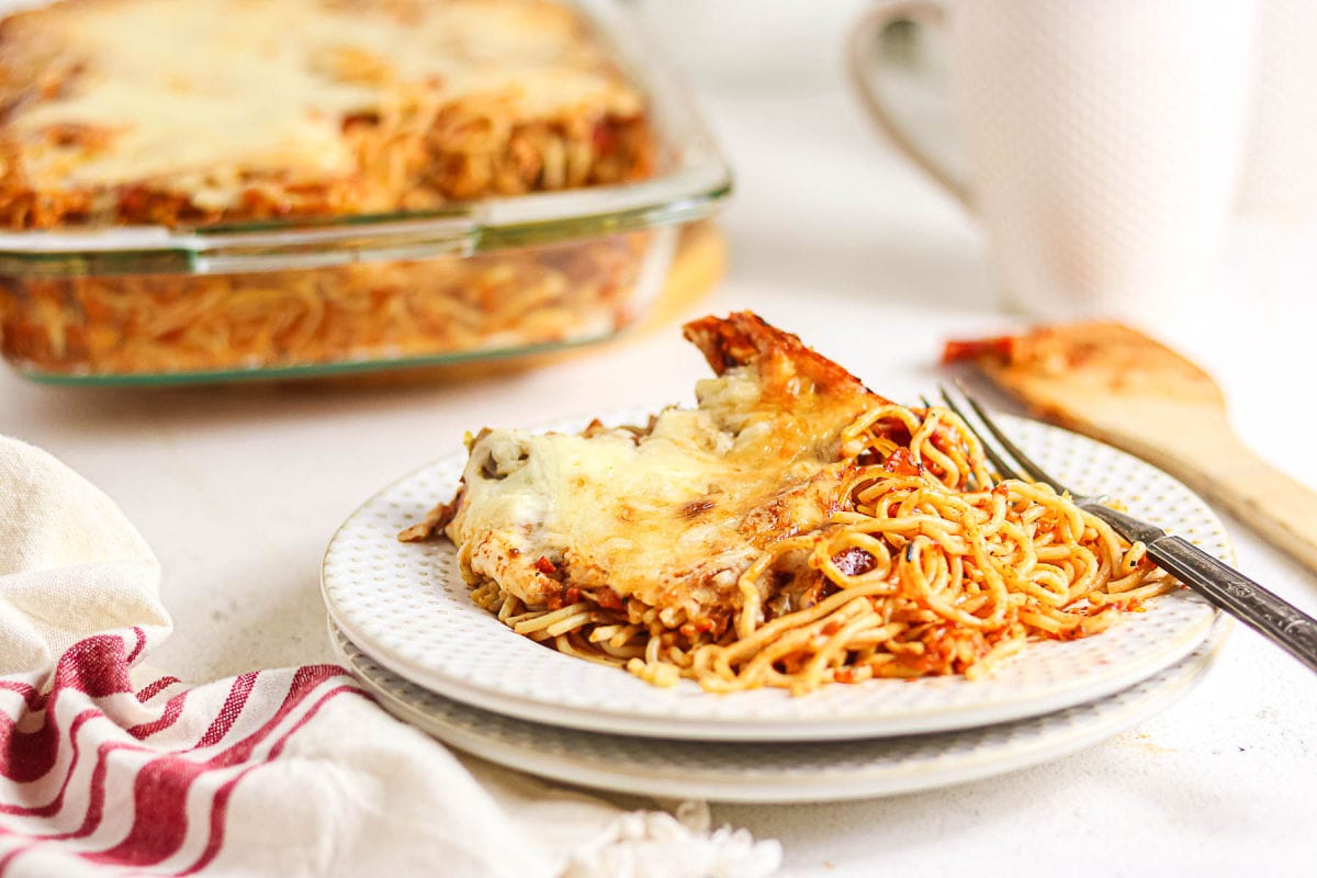 Serving of cheesy chicken spaghetti with red sauce on a plate with the casserole in the background