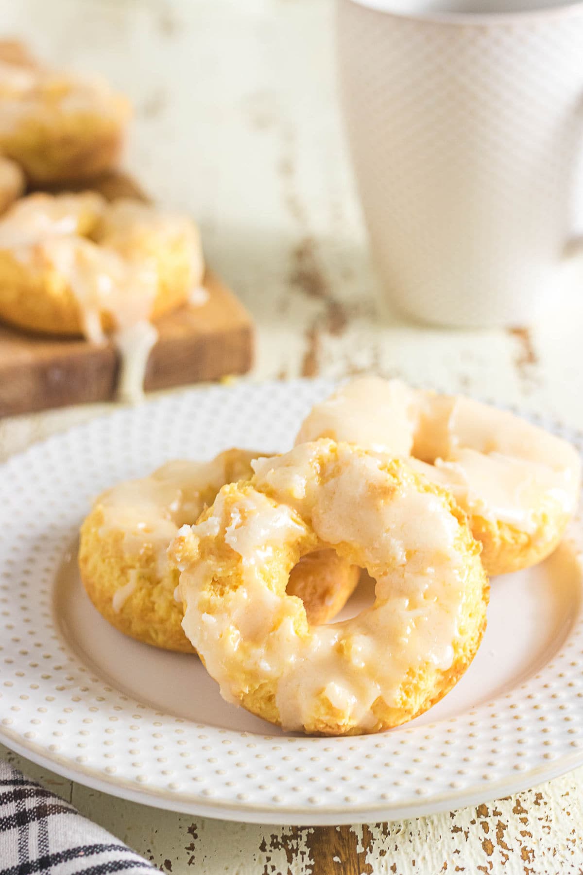 Three glazed sour cream baked donuts served on a white plate with a coffee mug in the background.