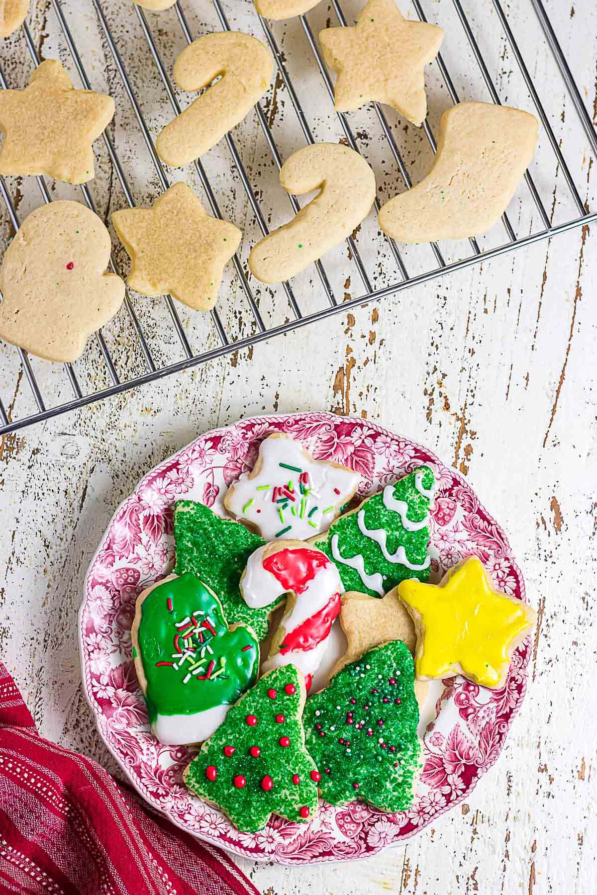 Plain Christmas sugar cookies cooling on a wire rack beside a plate of colorful decorated sugar cookies.