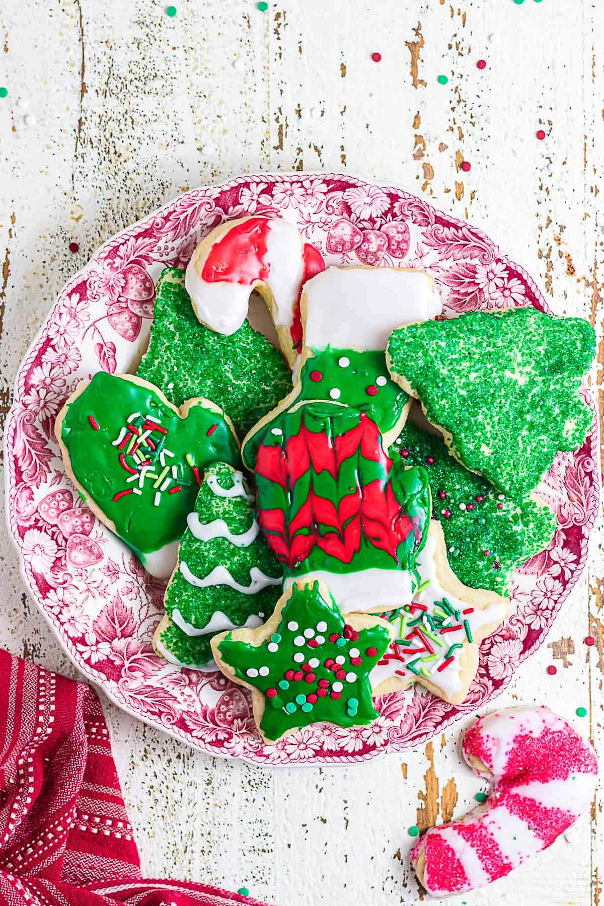 Homemade Christmas sugar cookies decorated with colorful icing, sparkling green sugar, and holiday sprinkles on a red and white plate.