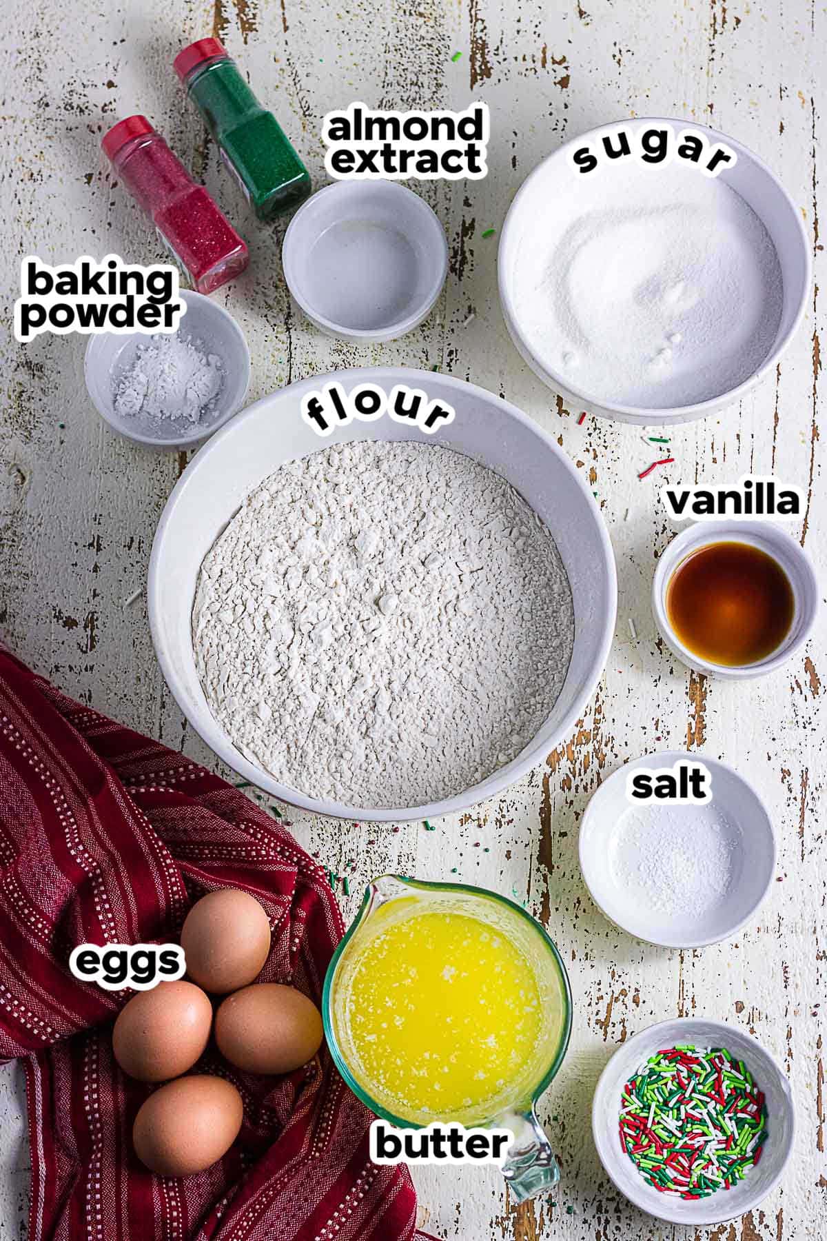 Ingredients for homemade Christmas sugar cookies including flour, sugar, butter, eggs, vanilla, almond extract, and baking powder.