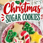 Christmas sugar cookies decorated with red, green, and white icing and festive sprinkles on a vintage red transferware plate, with plain cutout cookies on a cooling rack.