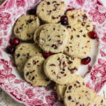 Homemade cranberry orange slice-and-bake cookies piled on a vintage red transferware plate with cranberries scattered around them.