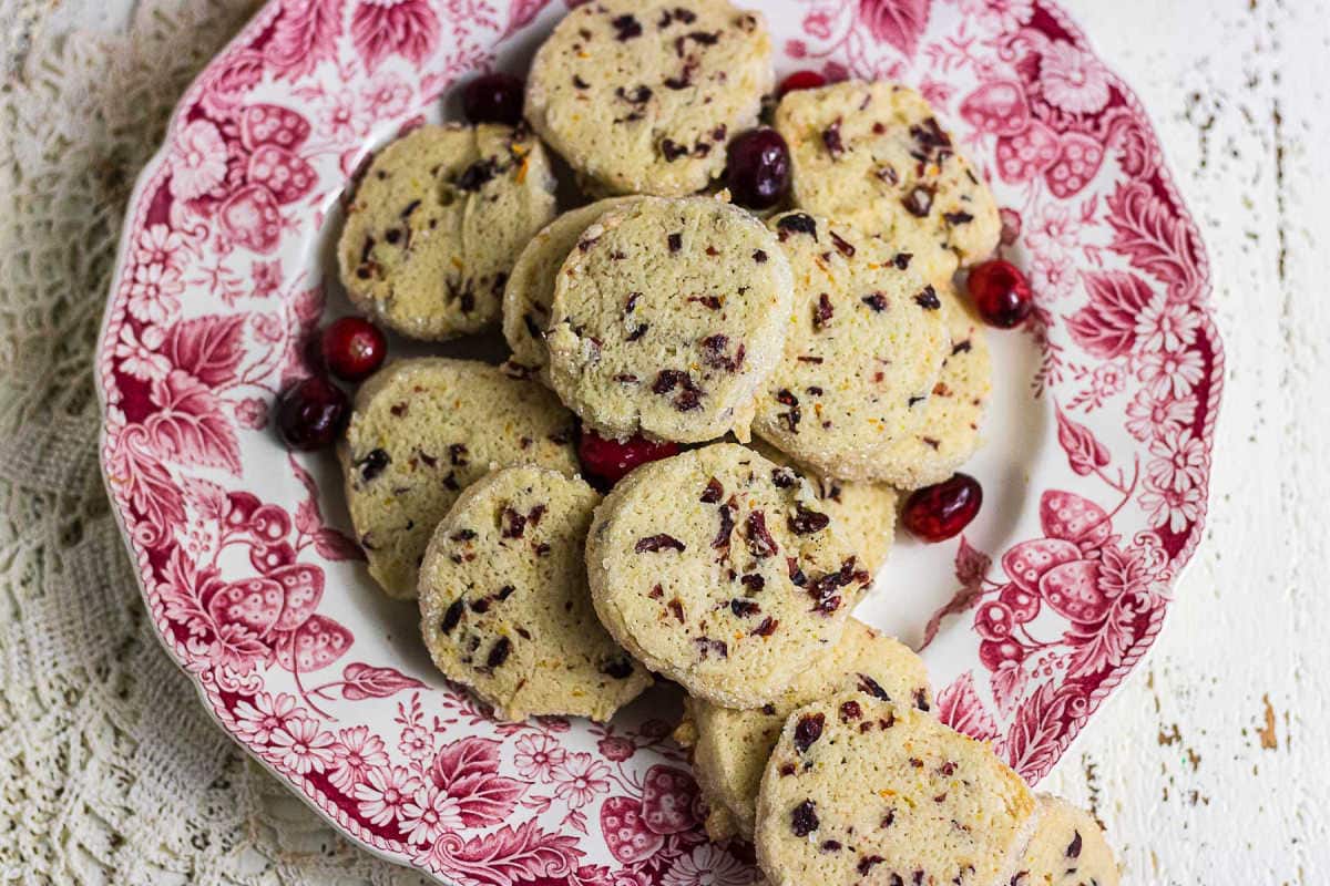 Homemade cranberry orange slice-and-bake cookies piled on a vintage red transferware plate with cranberries scattered around them.