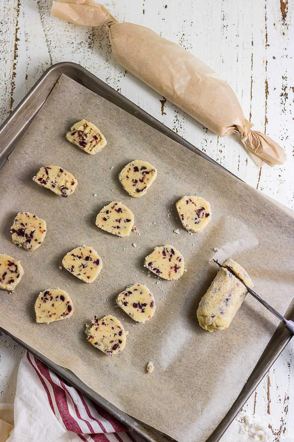 Sliced cranberry orange icebox cookie dough on a parchment-lined baking sheet beside a wrapped dough log and knife, ready for baking.