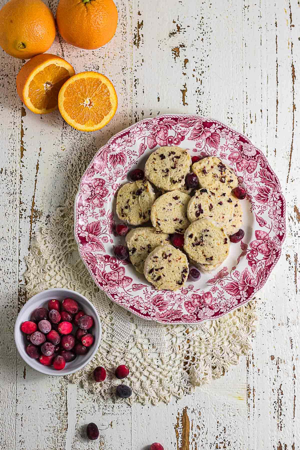 Overhead view of cranberry orange icebox cookies on a vintage red and white plate with fresh oranges, cranberries, and lace.